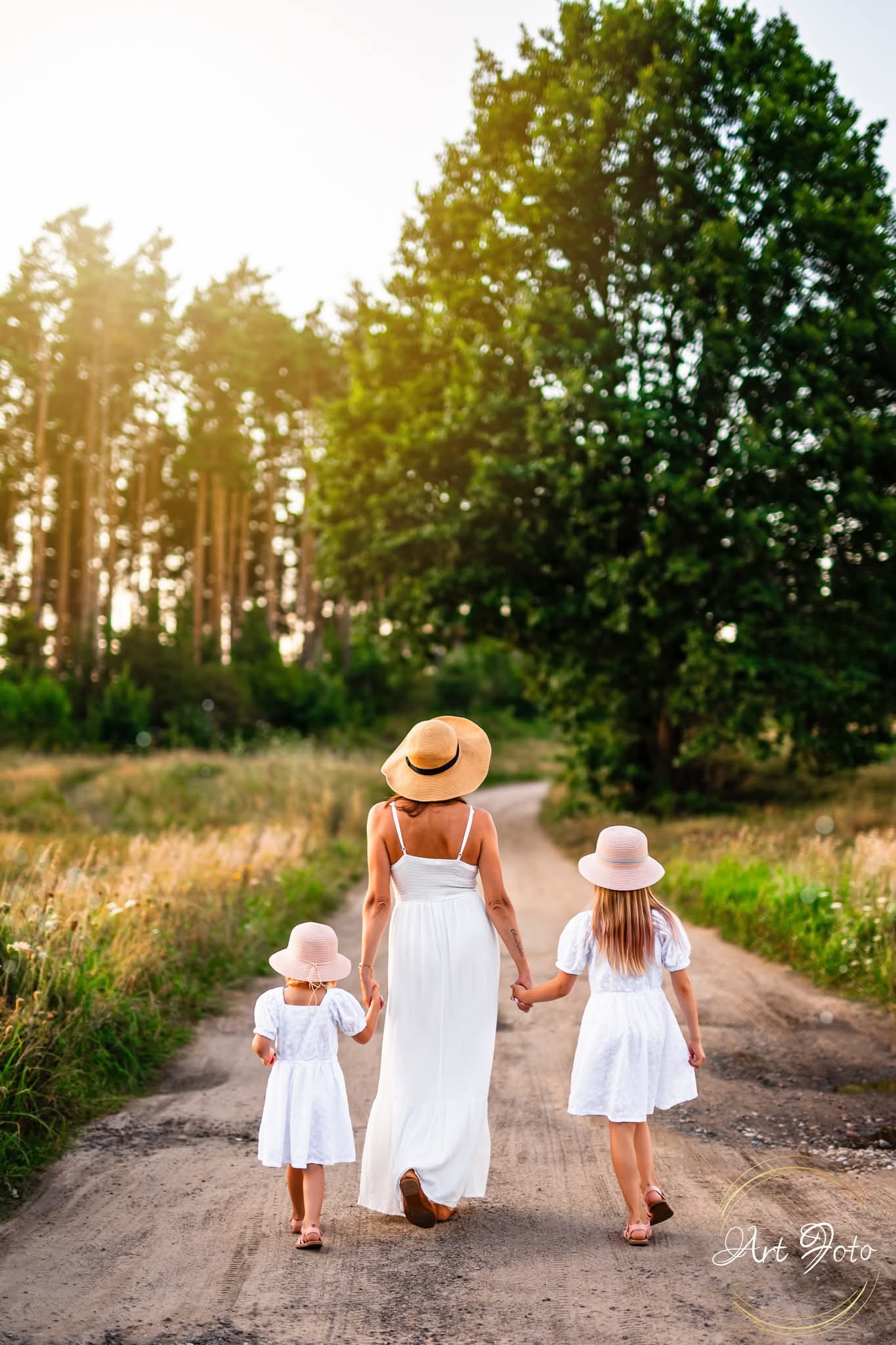Mama w kapeluszu i dwie córki w białych sukienkach idą polną drogą, trzymając się za ręce. Ciepłe światło zachodzącego słońca. Widok z tyłu.