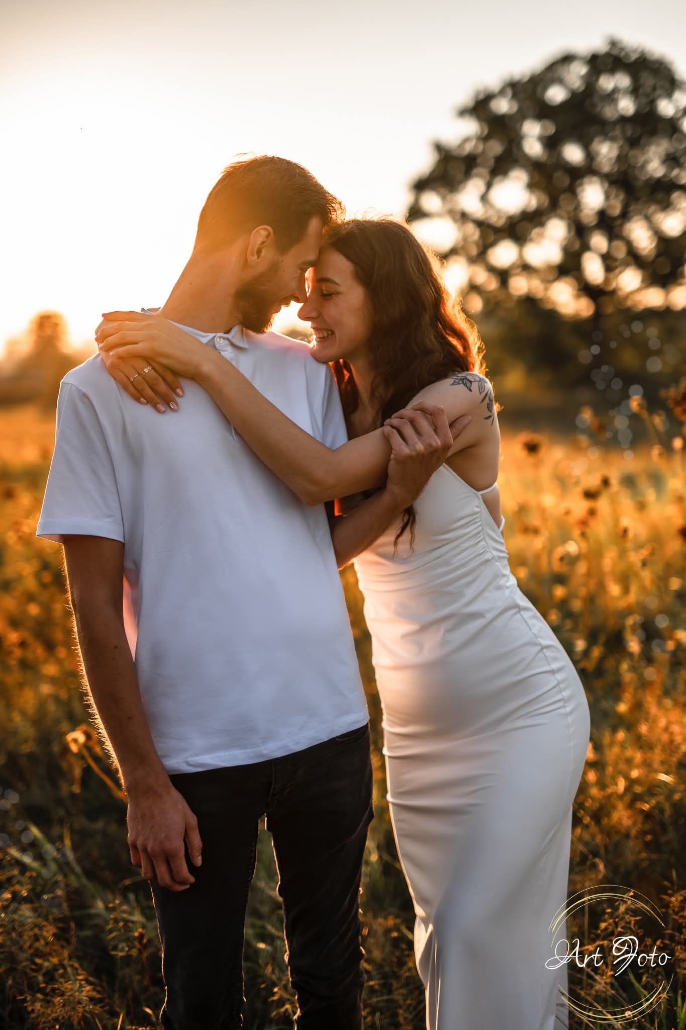Zakochana para w objęciach na tle łąki o zachodzie słońca. Kobieta w białej sukience, mężczyzna w białej koszulce polo. Delikatne światło podkreśla intymność chwili.