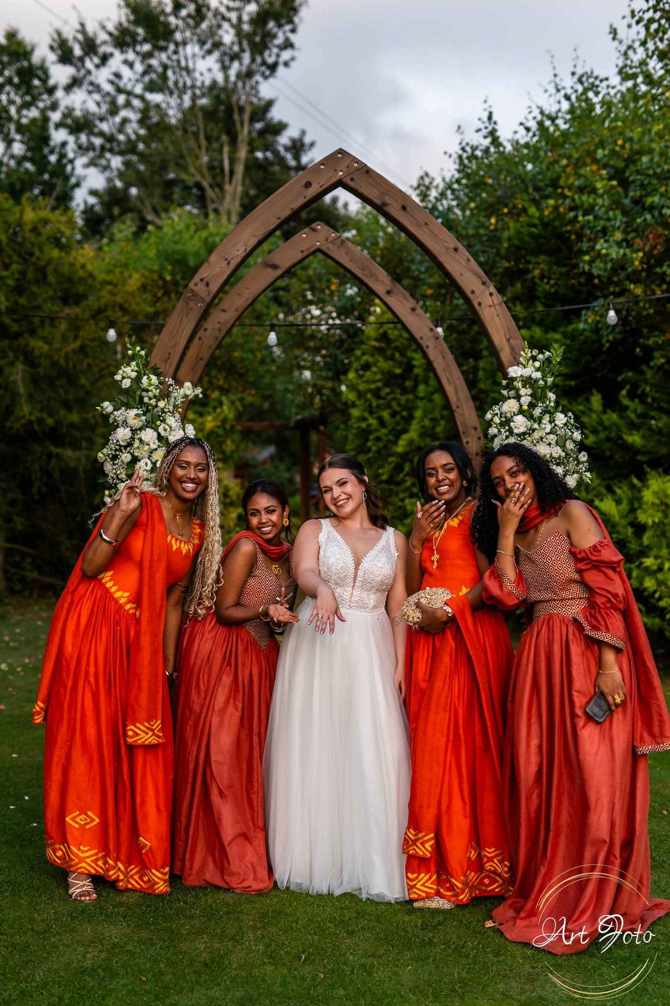 Panna młoda w białej sukni i druhny w pomarańczowych sukniach pozują pod drewnianym łukiem udekorowanym kwiatami. Zdjęcie wykonane w Gdyni.