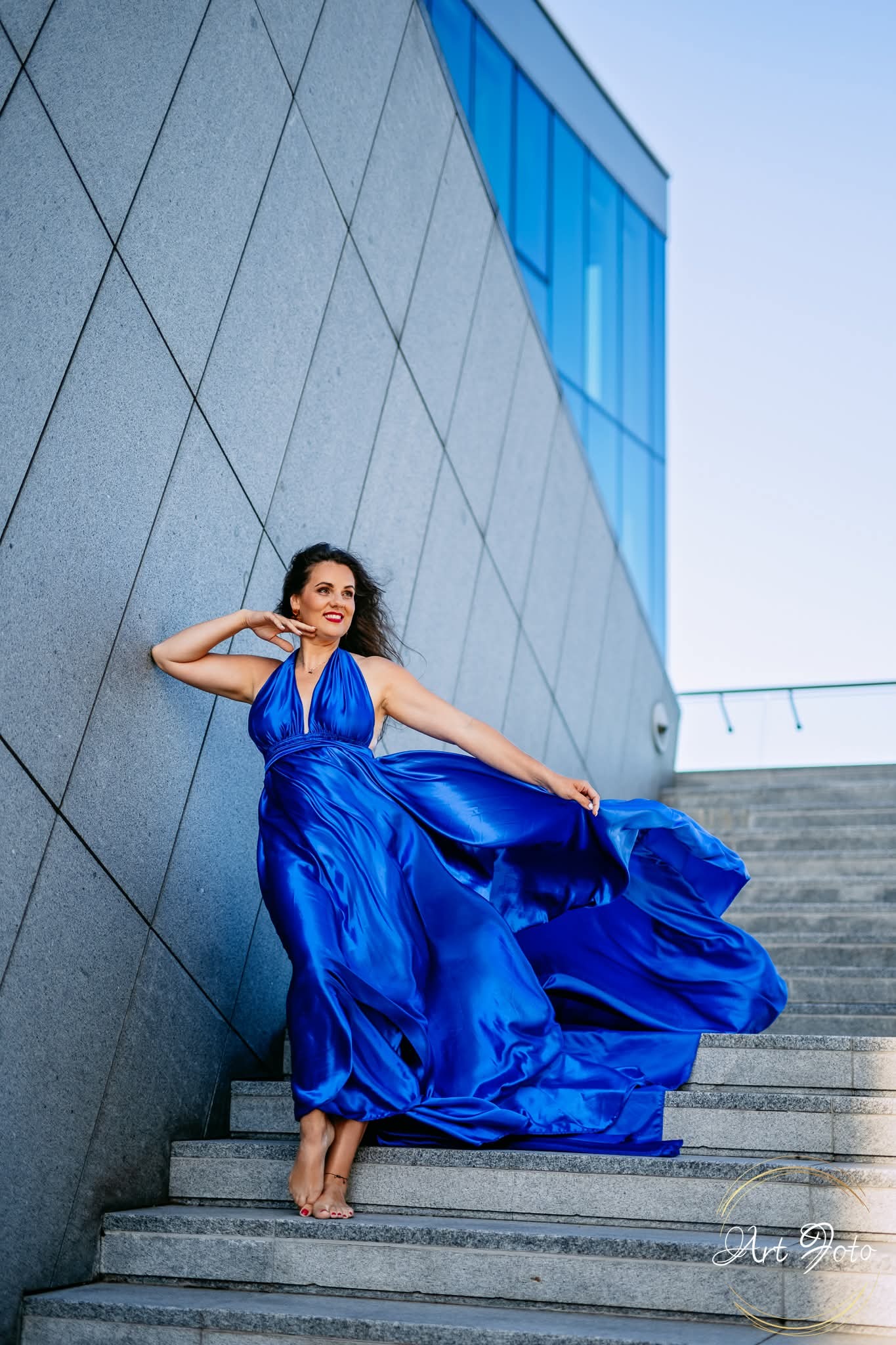Kobieta w powiewnej, błękitnej sukni pozuje na schodach nowoczesnego budynku. Dynamiczne ujęcie, podkreślone światłem i architekturą. Widoczny znak wodny fotografa.