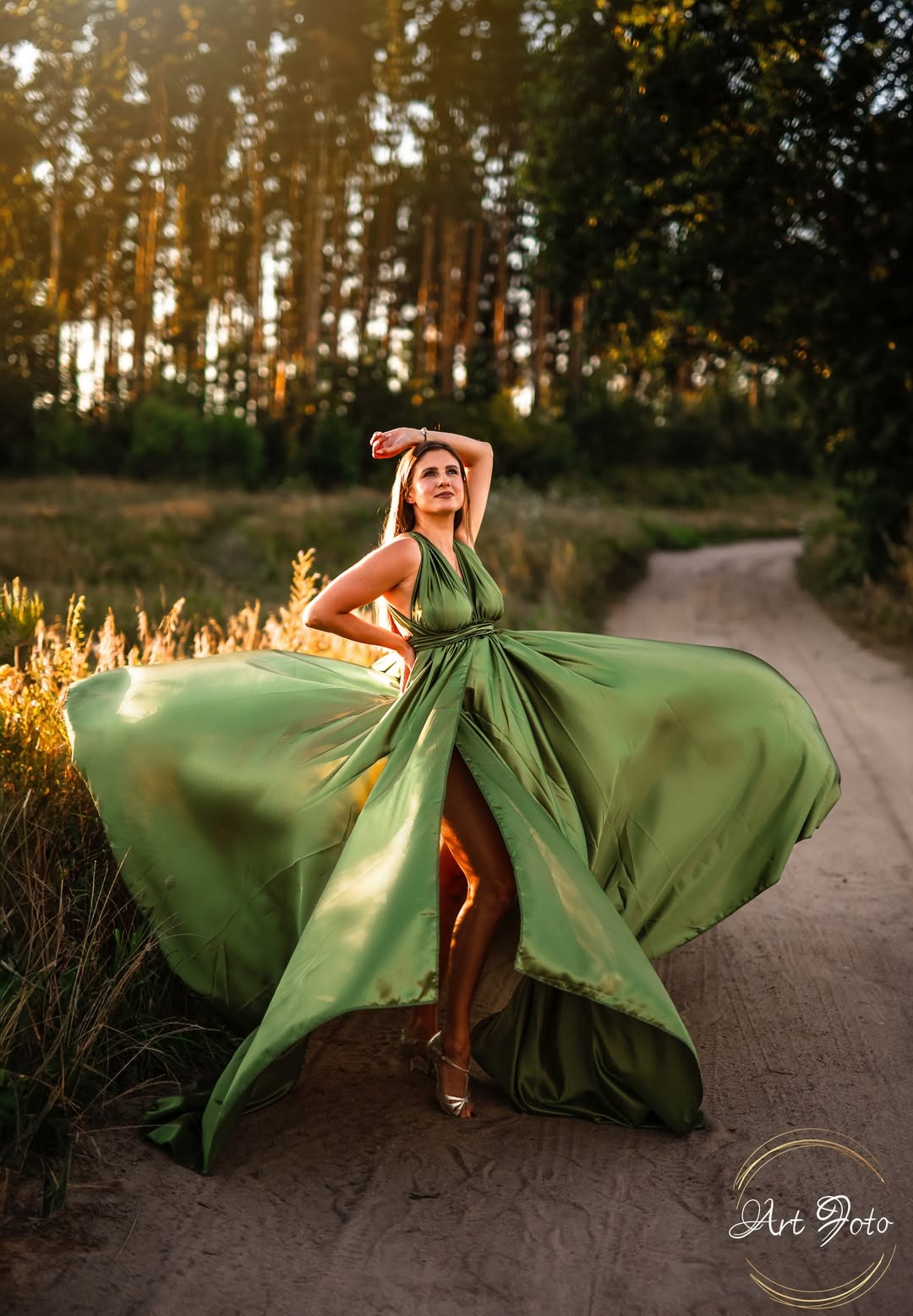 Kobieta w powiewnej, zielonej sukni na polnej drodze, pozuje w promieniach zachodzącego słońca. Widoczny las w tle i logo fotografa w prawym dolnym rogu.