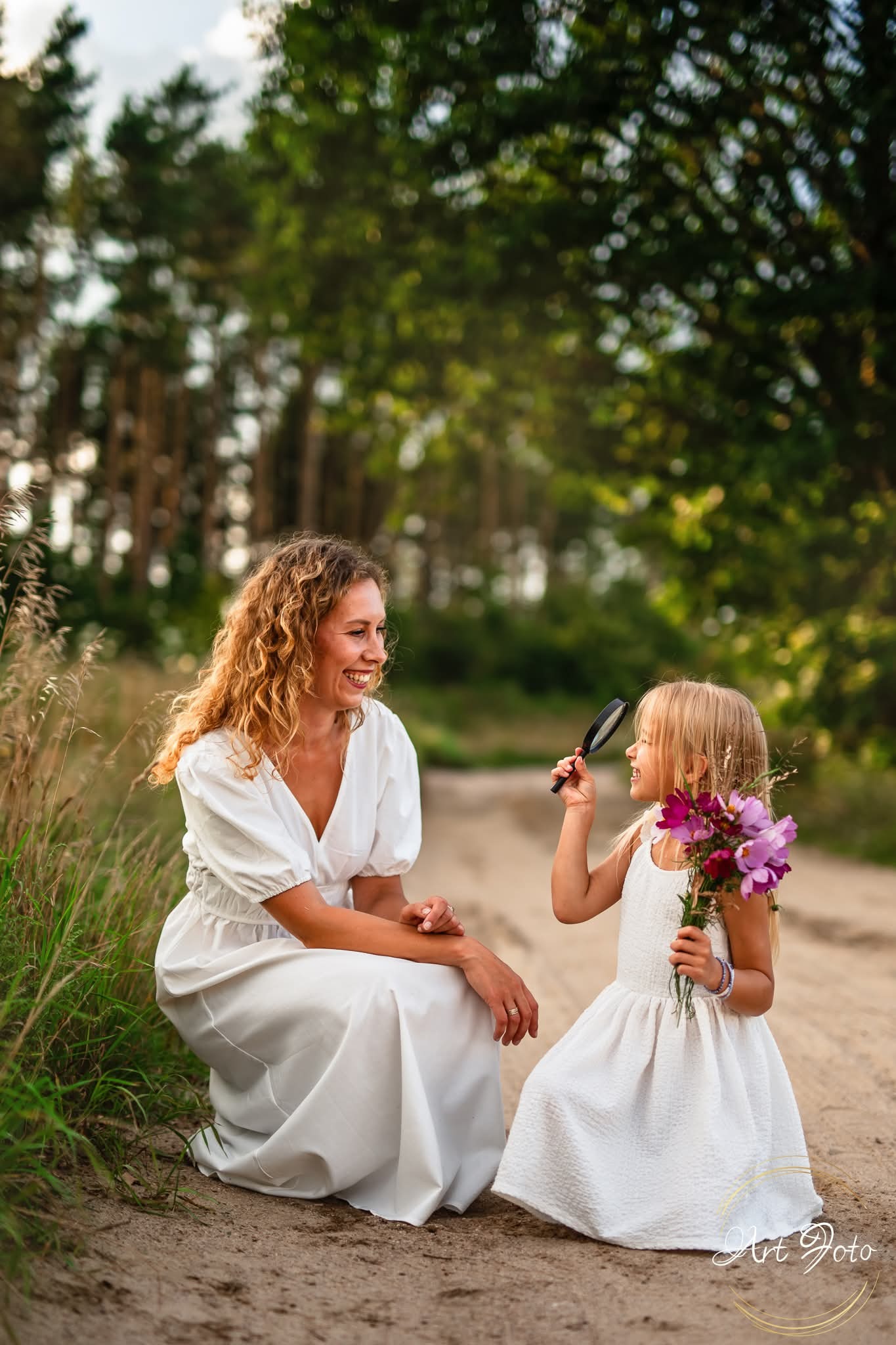 Sesja rodzinna w plenerze: uśmiechnięta mama w białej sukience kuca obok córki z lupą i bukietem fioletowych kwiatów na piaszczystej ścieżce.
