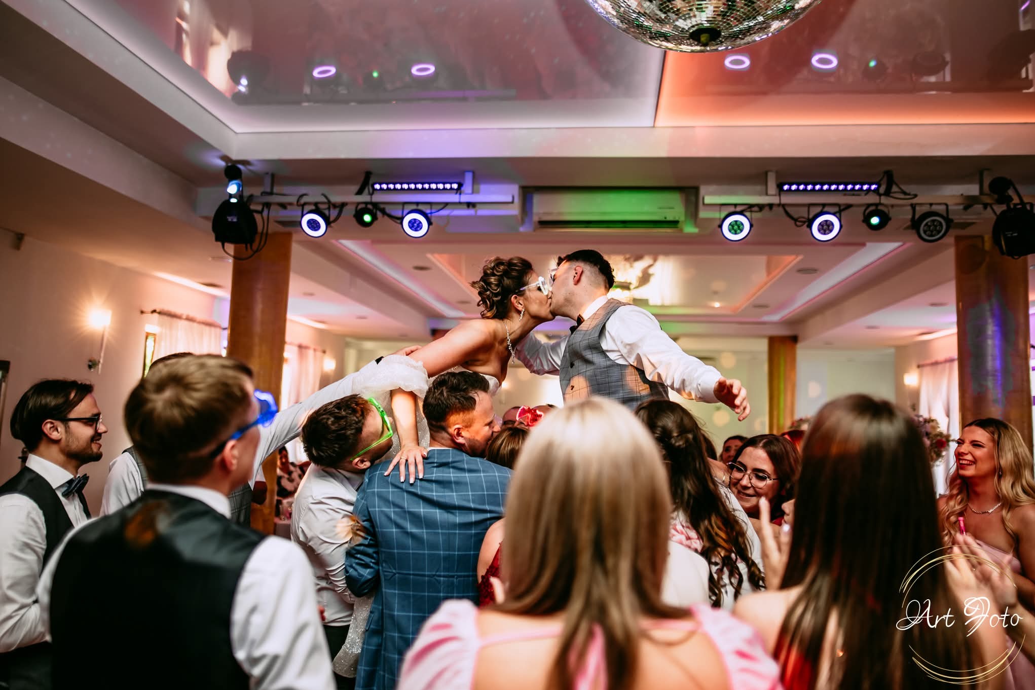 Panna młoda i pan młody w okularach, unoszeni na krzesłach przez gości, całują się na parkiecie podczas wesela. W tle oświetlenie dyskotekowe i kula lustrzana.