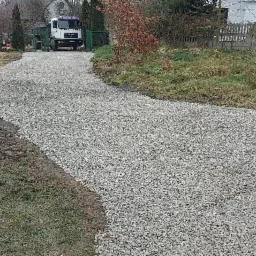 Nowo utwardzona droga z szarego kruszywa prowadząca do posesji, z widocznym samochodem ciężarowym w tle i stertą drewna po lewej stronie. Świeżo ułożona nawierzchnia kontrastuje z błotnistym...