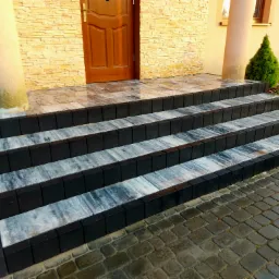 Nowe schody wejściowe z kostki brukowej o czarnych obrzeżach i szaro-białych płytach, prowadzące do drzwi wejściowych z dwoma kolumnami i elewacją z kamienia dekoracyjnego.