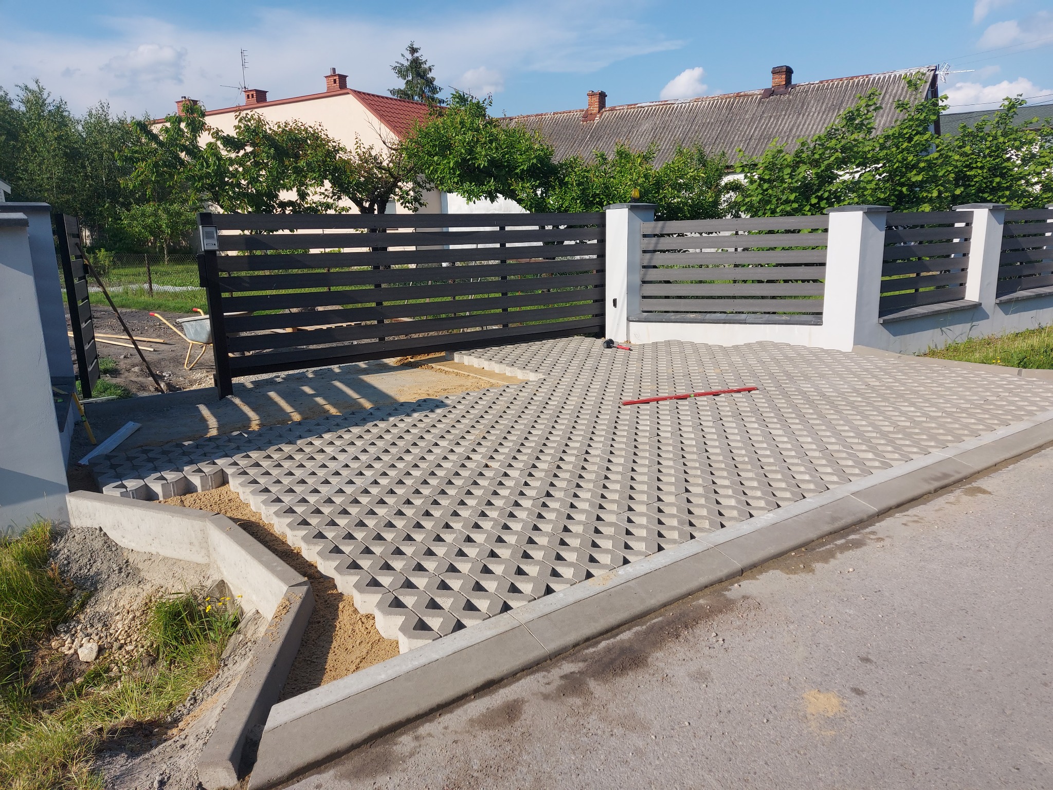 Układanie ażurowej kostki brukowej na podjeździe, widoczny poziomujący laser oraz brama w tle.