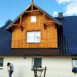 Dom z dobudowaną drewnianą nadbudówką z oknami, niebieski dach z dachówki, mężczyzna zamiatający przed posesją.