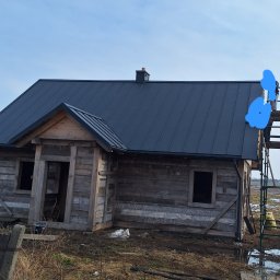 Bogacki sp. z o.o. - Surowy, drewniany dom z ciemnym, metalowym dachem w trakcie budowy. Widoczne rusztowania i elementy wykończeniowe. Krajobraz wiejski, pochmurne niebo.