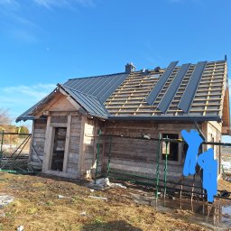 Bogacki sp. z o.o. - Drewniany dom w trakcie krycia dachu blachą na rąbek stojący. Widoczne rusztowania, więźba dachowa i częściowo ułożone panele blachy w kolorze grafitowym.