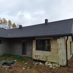 Bogacki sp. z o.o. - Surowy stan domu z szarego bloczka i ciemnym dachem z dachówki. Widoczne okna, rynna i komin. Otoczenie zarośnięte, z resztkami materiałów budowlanych.