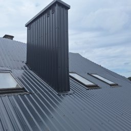 Bogacki sp. z o.o. - Szary, blaszany dach z kominem wentylacyjnym i oknami dachowymi, widok z góry, pochmurne niebo w tle. Charakterystyczne pionowe żłobienia blachy.