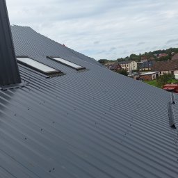 Bogacki sp. z o.o. - Szary dach z blachy trapezowej z dwoma oknami dachowymi, widok z góry, w tle zabudowa mieszkalna i pochmurne niebo.