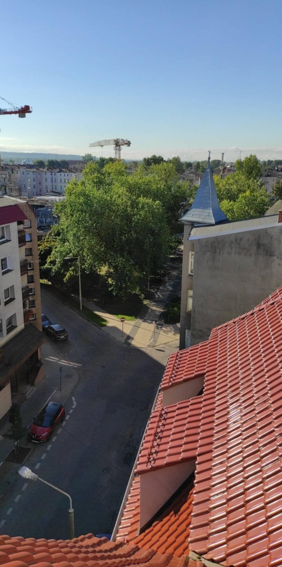 Widok z góry na czerwoną dachówkę, ulicę z samochodami i panoramę miasta z dźwigami budowlanymi w tle.