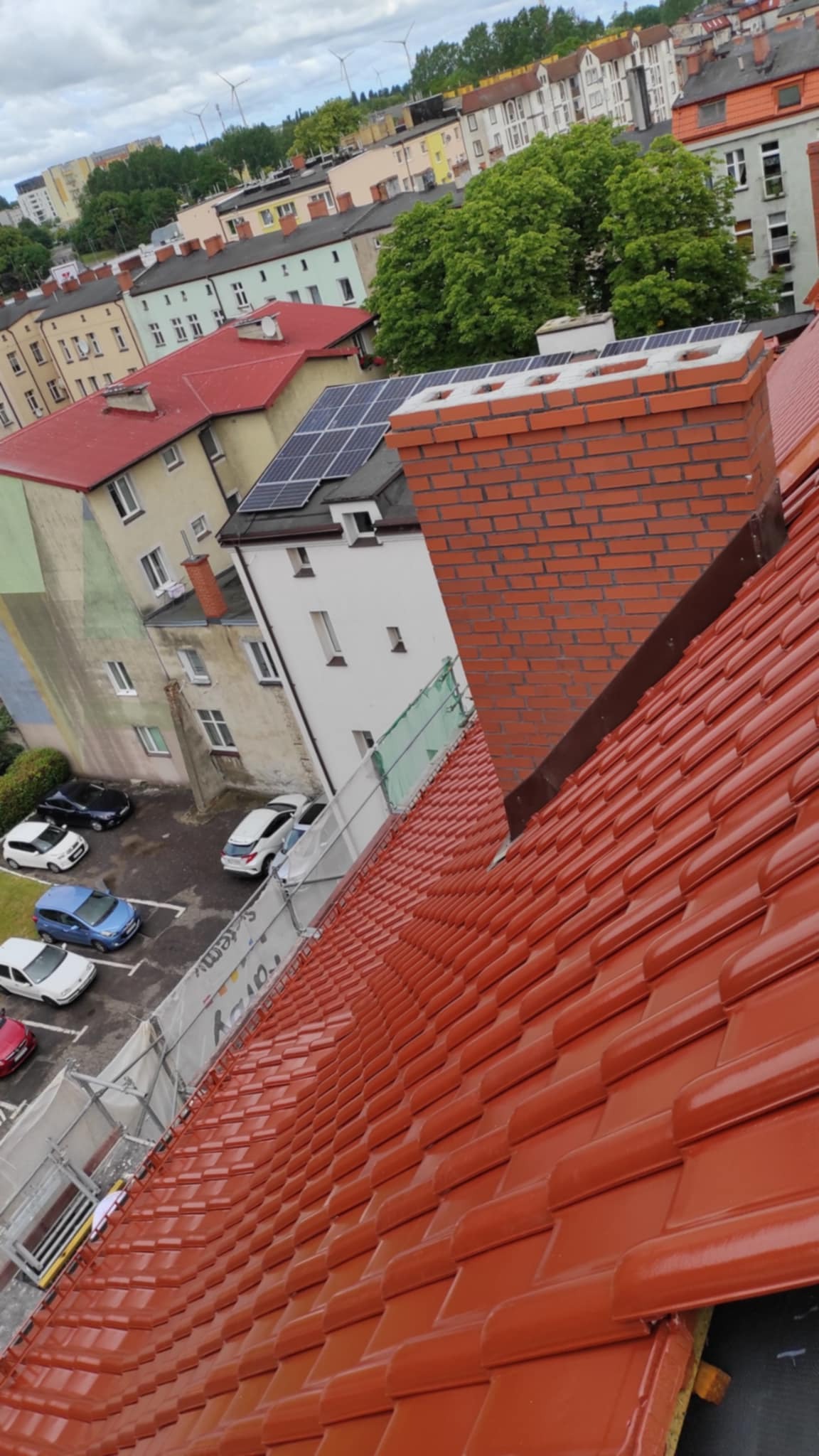 Nowy, lśniący dach z czerwonej dachówki ceramicznej, widok z góry na komin z cegły klinkierowej, panele słoneczne na sąsiednim budynku i parking z autami w tle.