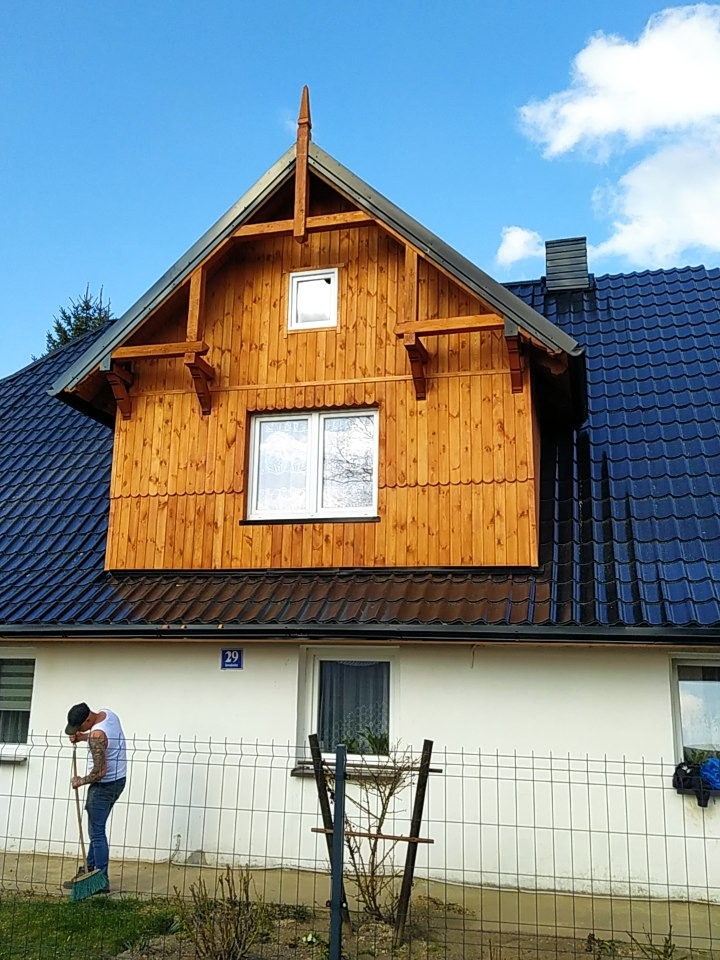 Dom z dobudowaną drewnianą nadbudówką z oknami, niebieski dach z dachówki, mężczyzna zamiatający przed posesją.