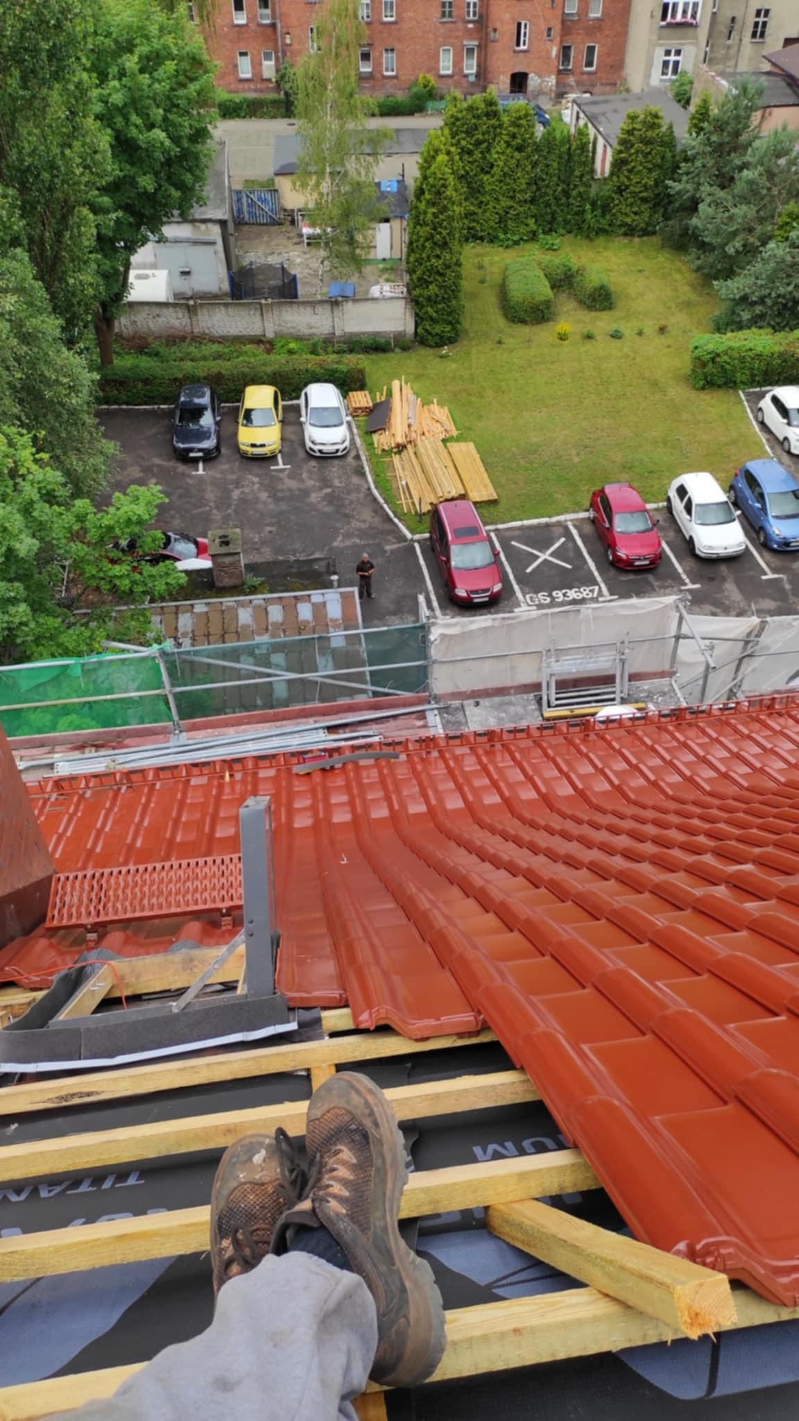 Widok z góry na częściowo pokryty, błyszczący, czerwony dach z widocznymi łatami i membraną, w tle parking z samochodami i budynki mieszkalne.