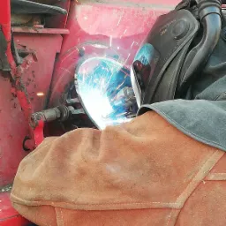 Spawanie elementu czerwonej maszyny rolniczej z użyciem maski spawalniczej 3M Speedglas 9100 FX i ochronnego skórzanego rękawa.