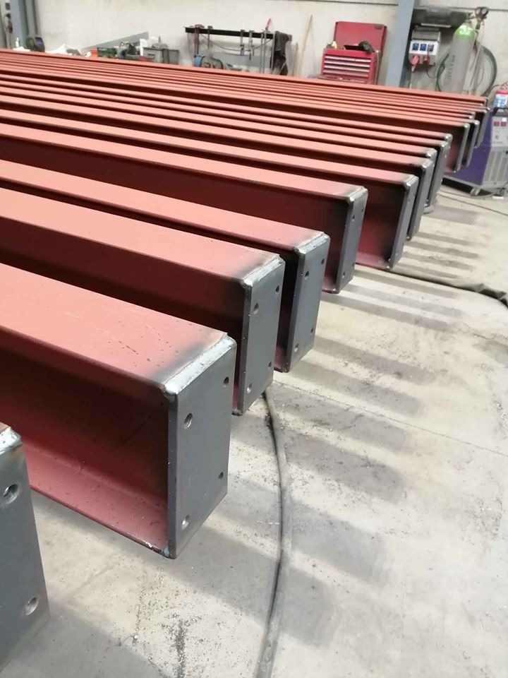 Rząd czerwonych, stalowych belek o przekroju ceownika, ustawionych równolegle, z szarymi, spawane końcówkami z otworami na śruby, leżących na betonowej podłodze w warsztacie.