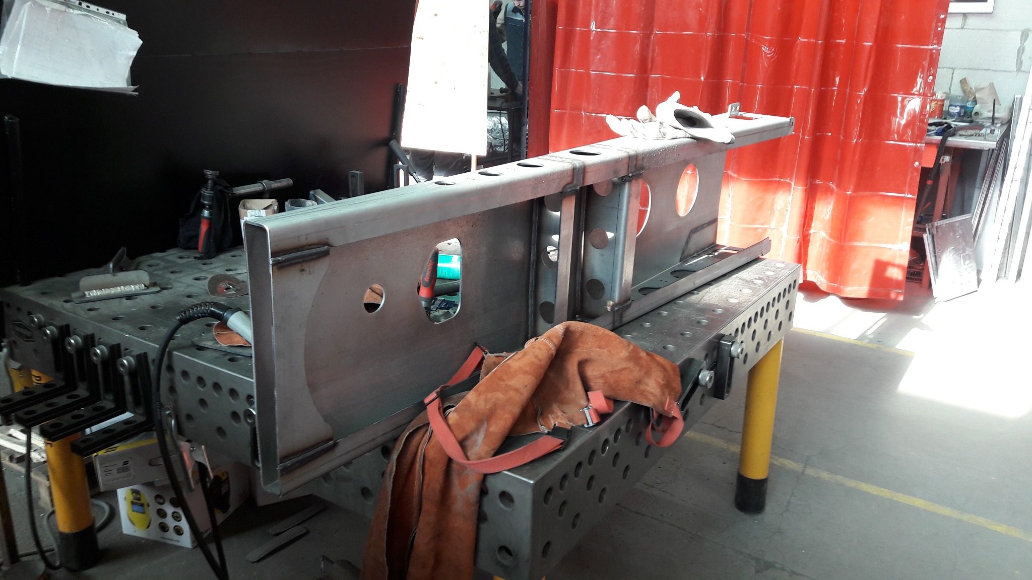 Metalowa konstrukcja z otworami na perforowanym stole spawalniczym, z widocznymi śladami spawania i elementami mocującymi. Ochronny fartuch skórzany leży obok, a w tle czerwona kurtyna spawalnicza.