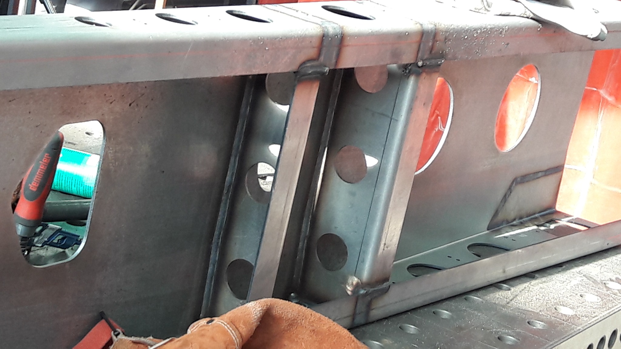 Metalowa konstrukcja z otworami, połączona spawami, widoczne narzędzie Demmeler i rękawica ochronna.