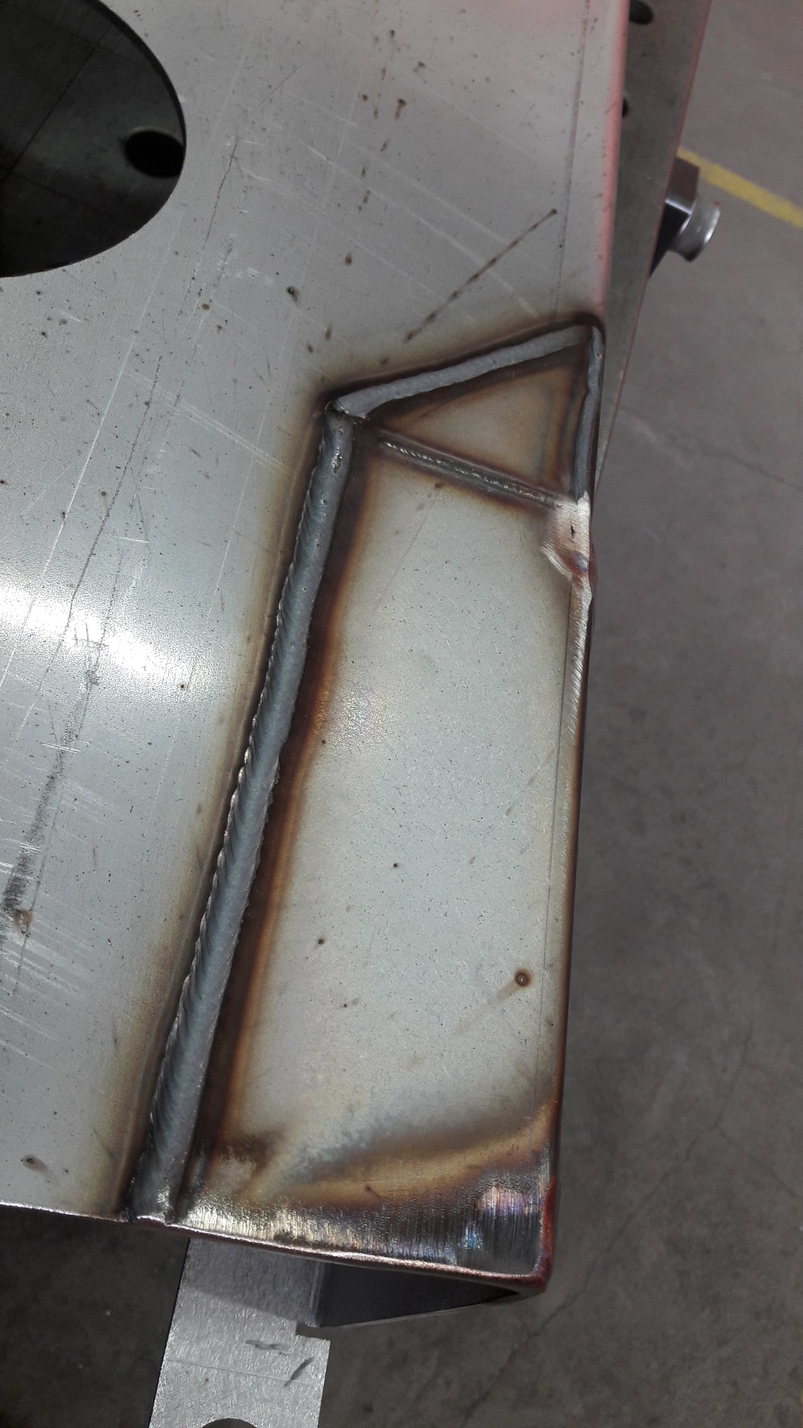 Spoina narożna na metalowej płycie, widoczne przebarwienia termiczne i delikatne zarysowania na powierzchni, ślad po cięciu na krawędzi.