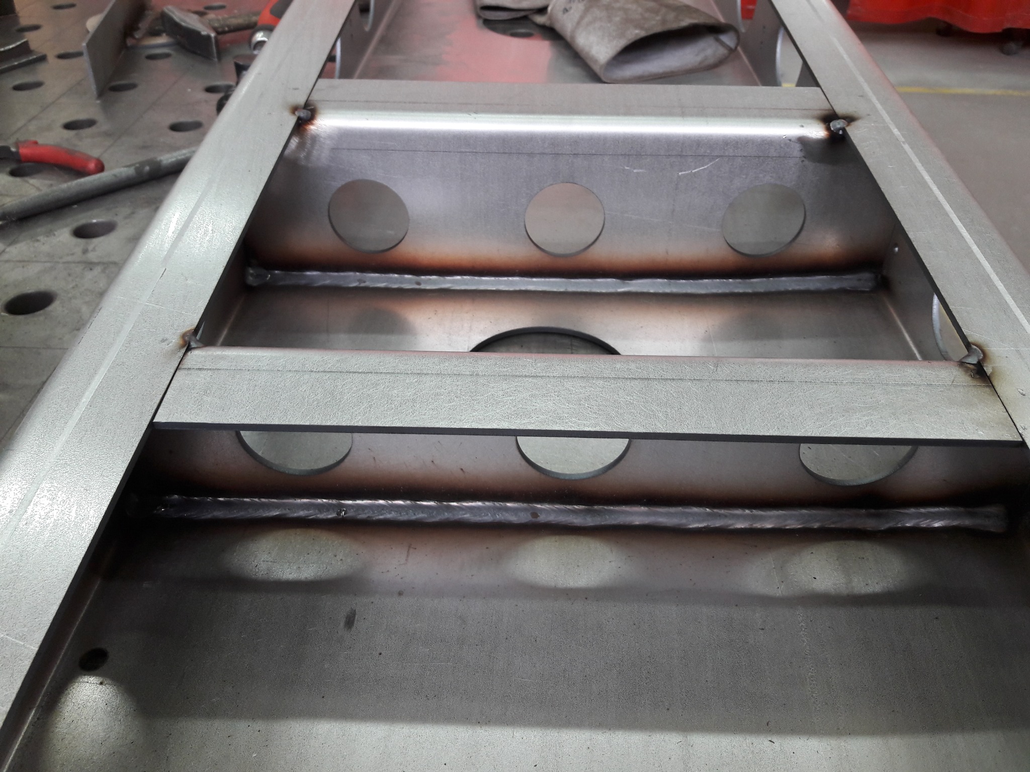 Metalowa konstrukcja skrzynkowa z otworami, spawana, widoczne spoiny po spawaniu, na stole warsztatowym, z narzędziami w tle.