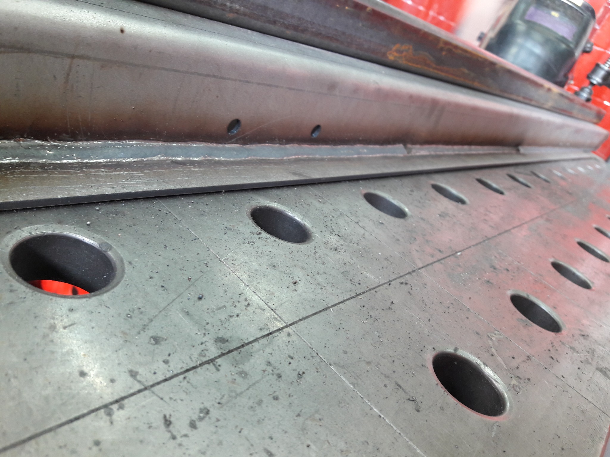 Stół spawalniczy z otworami i zamocowanym profilem, widoczna spawarka w tle.