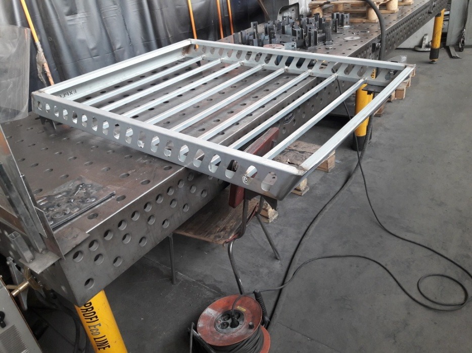 Stalowa konstrukcja kratowa z otworami na obwodzie, spawana na stole spawalniczym z widocznymi narzędziami i przewodami w tle.