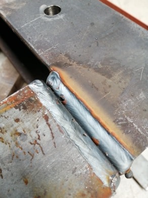 Spoina łącząca dwa elementy metalowe pod kątem prostym, widoczne przypalenia i nierówności spoiny, na górnym elemencie otwór.