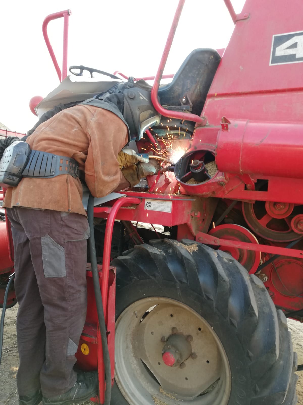 Pracownik w kombinezonie ochronnym spawa uszkodzony element czerwonego kombajnu rolniczego, iskry oświetlają miejsce naprawy.