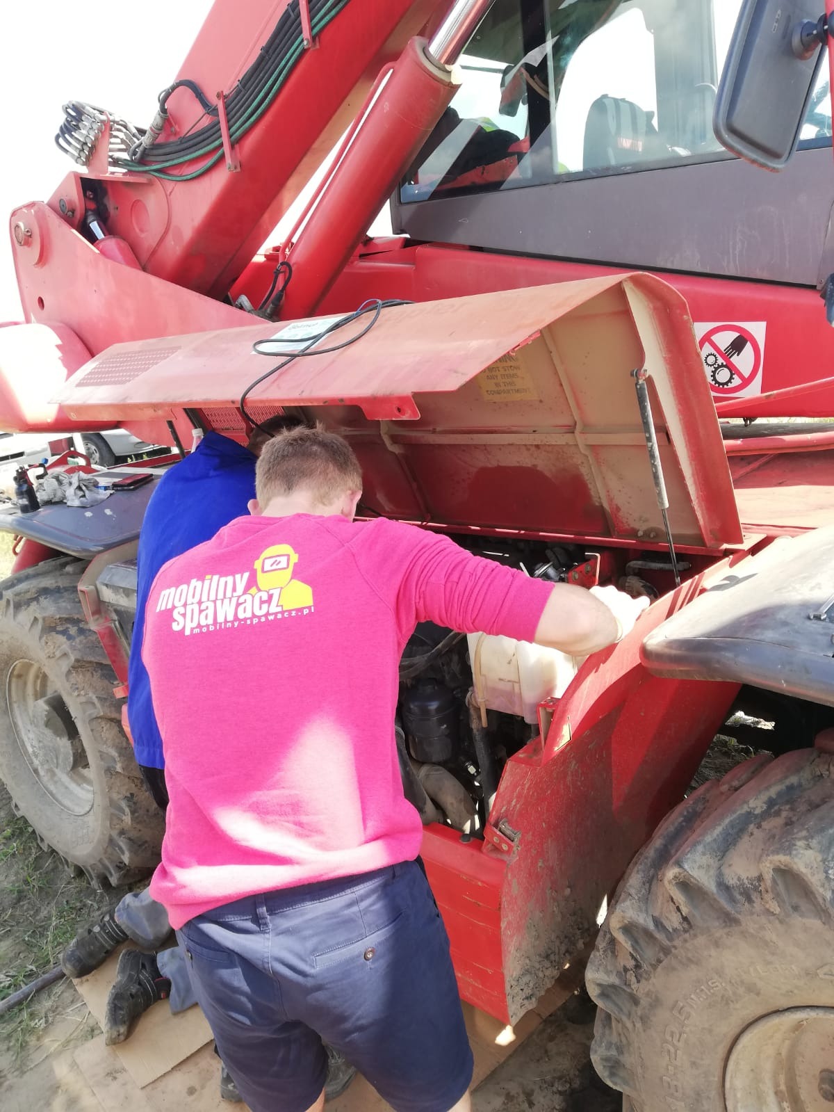 Dwóch mężczyzn w trakcie naprawy silnika czerwonego wózka widłowego na zewnątrz, jeden z nich w różowej bluzie z logo firmy.