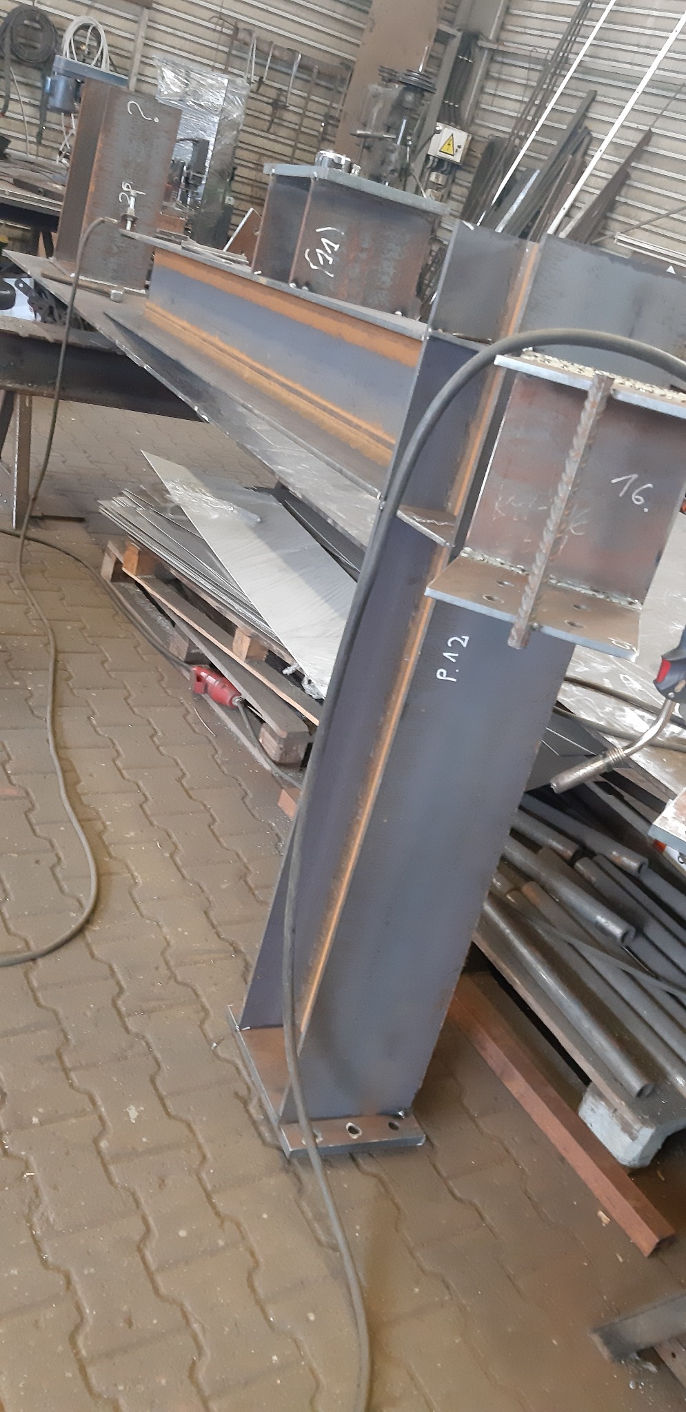 Fragment konstrukcji stalowej z profili, spawana, z widocznymi oznaczeniami P.12 i 16, w warsztacie, na tle podłogi z kostki brukowej.