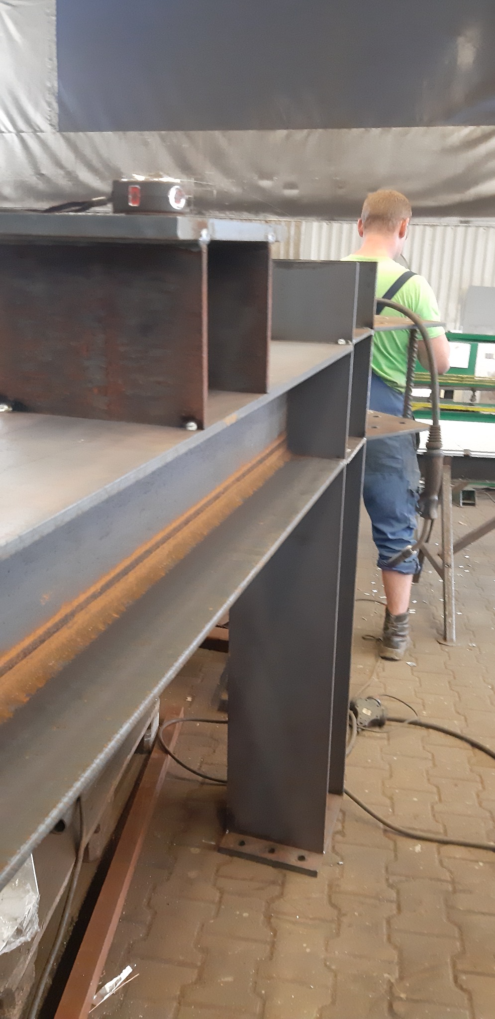 Konstrukcja stalowa z profili zamkniętych i dwuteowników, częściowo zardzewiała, spawana, w warsztacie, w tle pracownik w zielonej koszulce i niebieskich ogrodniczkach.