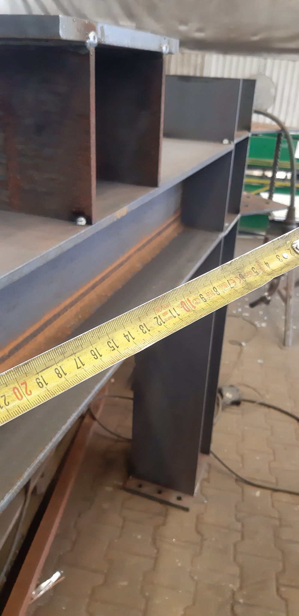 Metalowa konstrukcja w trakcie pomiaru, widoczna taśma miernicza z zaznaczonymi centymetrami, surowy stan wykończenia, spawane elementy.