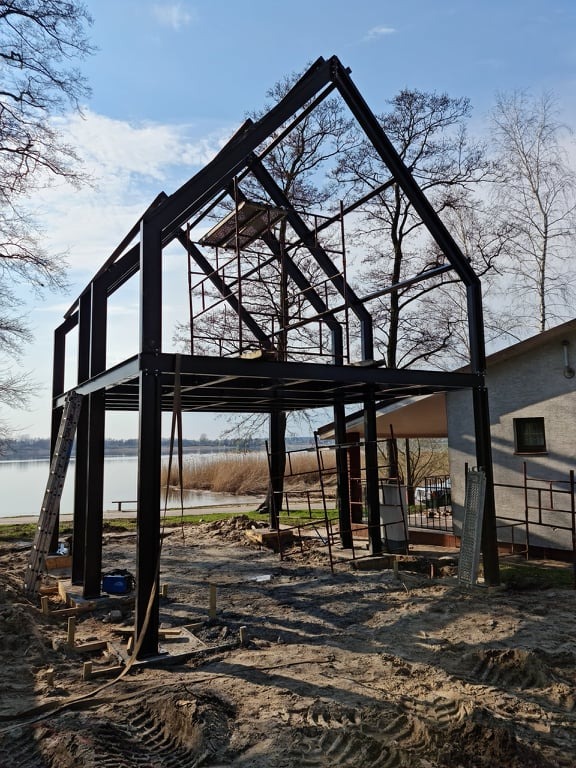 Surowa, dwukondygnacyjna konstrukcja szkieletowa domu z metalu w trakcie budowy, widoczna drabina i rusztowanie, tło stanowi jezioro i budynek w oddali.