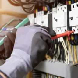 Elektryk w rękawicach ochronnych mierzy napięcie na stycznikach w rozdzielnicy elektrycznej za pomocą multimetru z przewodami pomiarowymi.