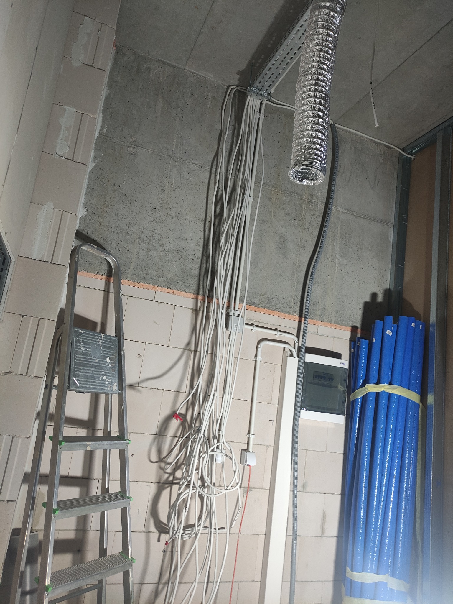 Instalacja elektryczna w surowym stanie: wiązka przewodów zwisająca z sufitu, aluminiowa rura wentylacyjna, rozdzielnia elektryczna, drabina aluminiowa i niebieskie rury PEX czekające na montaż...