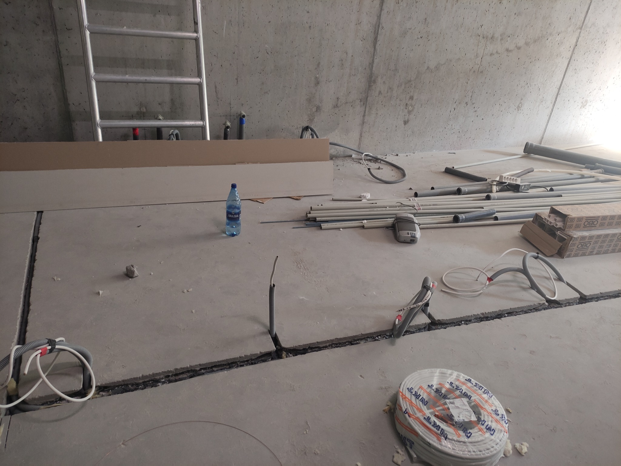 Surowy stan podłogi w trakcie prac elektrycznych: widoczne kable w peszlach w bruzdach, rury instalacyjne, skrzynki OBO, butelka wody i drabina oparta o betonową ścianę.