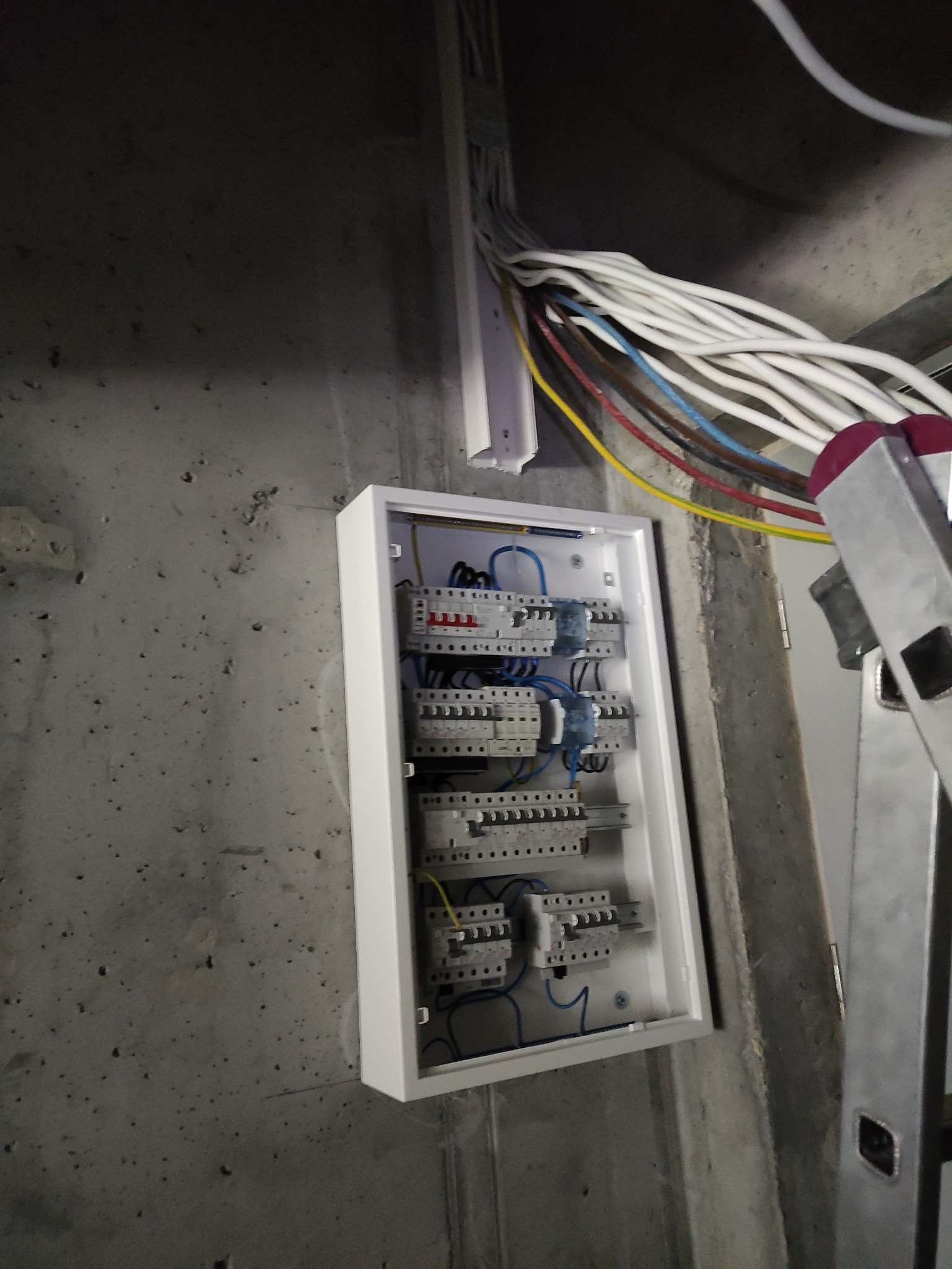Skrzynka z zabezpieczeniami elektrycznymi zamontowana na surowej ścianie w Warszawie, widoczne kable i drabina oparta o ścianę.