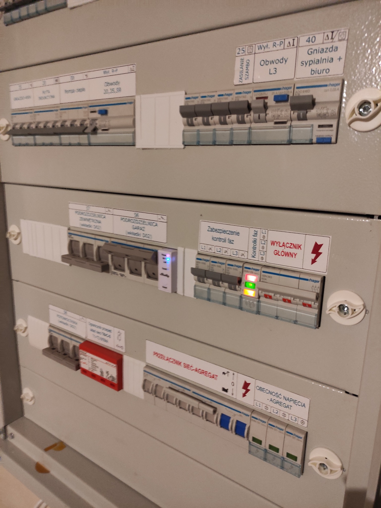 Rozdzielnica elektryczna Hager z widocznymi bezpiecznikami, wyłącznikami i kontrolkami faz. Elementy opisane etykietami, w tym 'Wyłącznik Główny' i 'Obecność Napięcia - Agregat'.