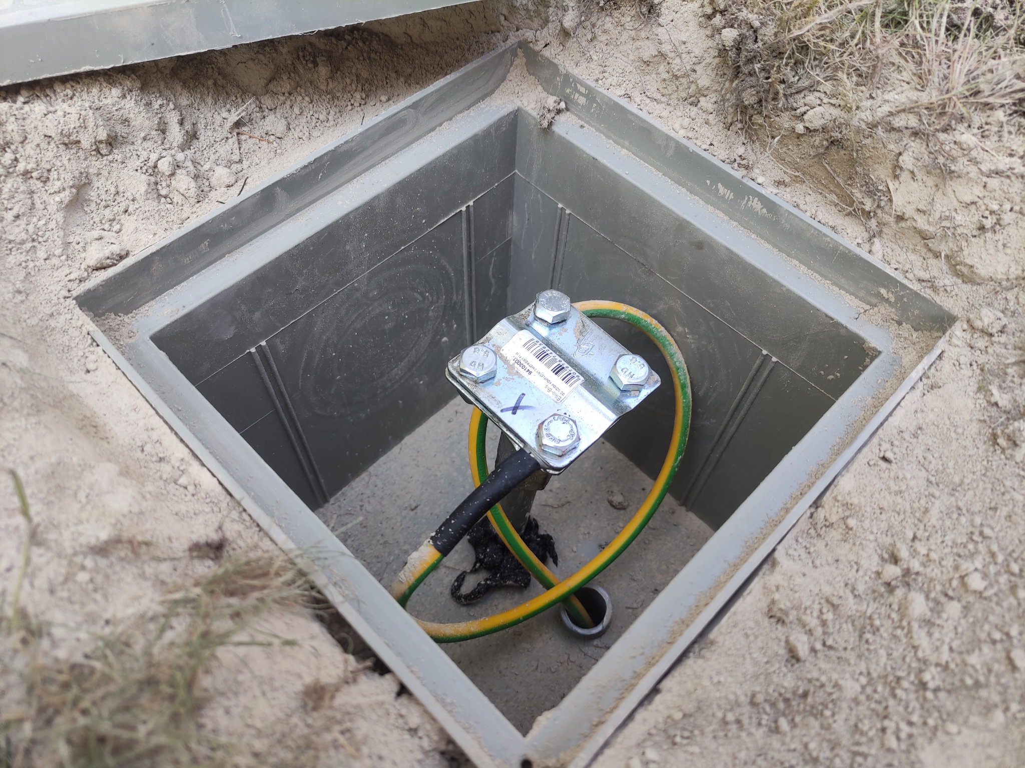 Uziemienie w studzience rewizyjnej z żółto-zielonym przewodem ochronnym i metalową płytką z naklejką, widoczne ślady ziemi, ujęcie z góry.