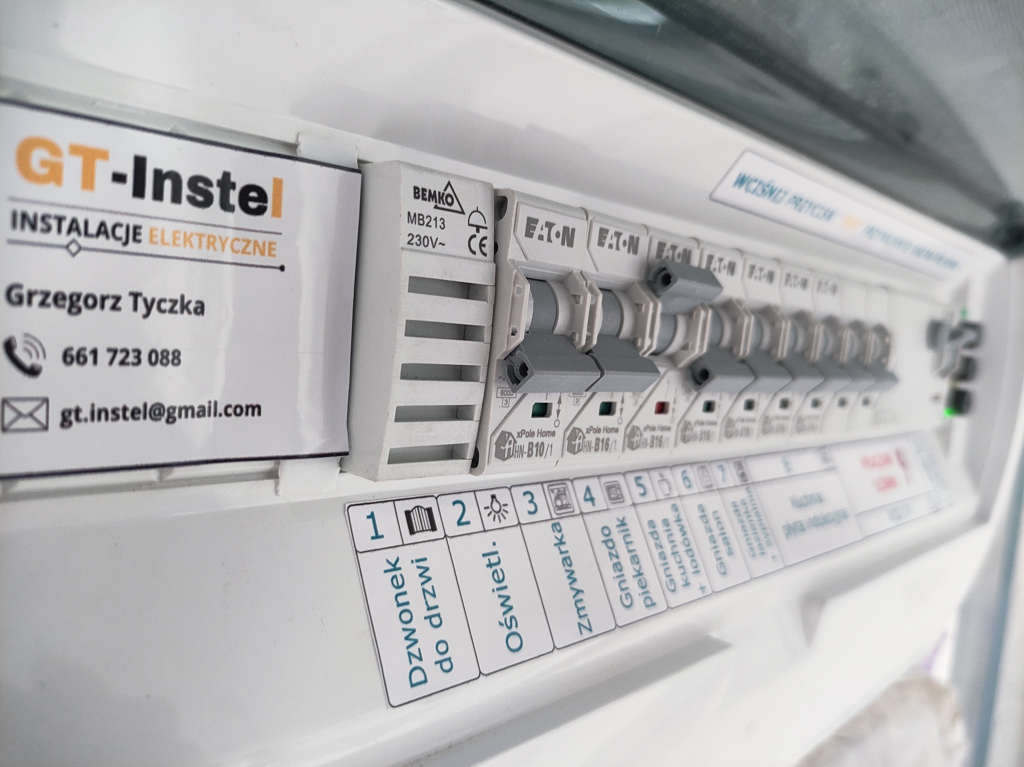 Rozdzielnica elektryczna z opisami obwodów, widoczne bezpieczniki Eaton i etykieta firmy GT-Instel z danymi kontaktowymi. Detal instalacji elektrycznej wewnątrz budynku.