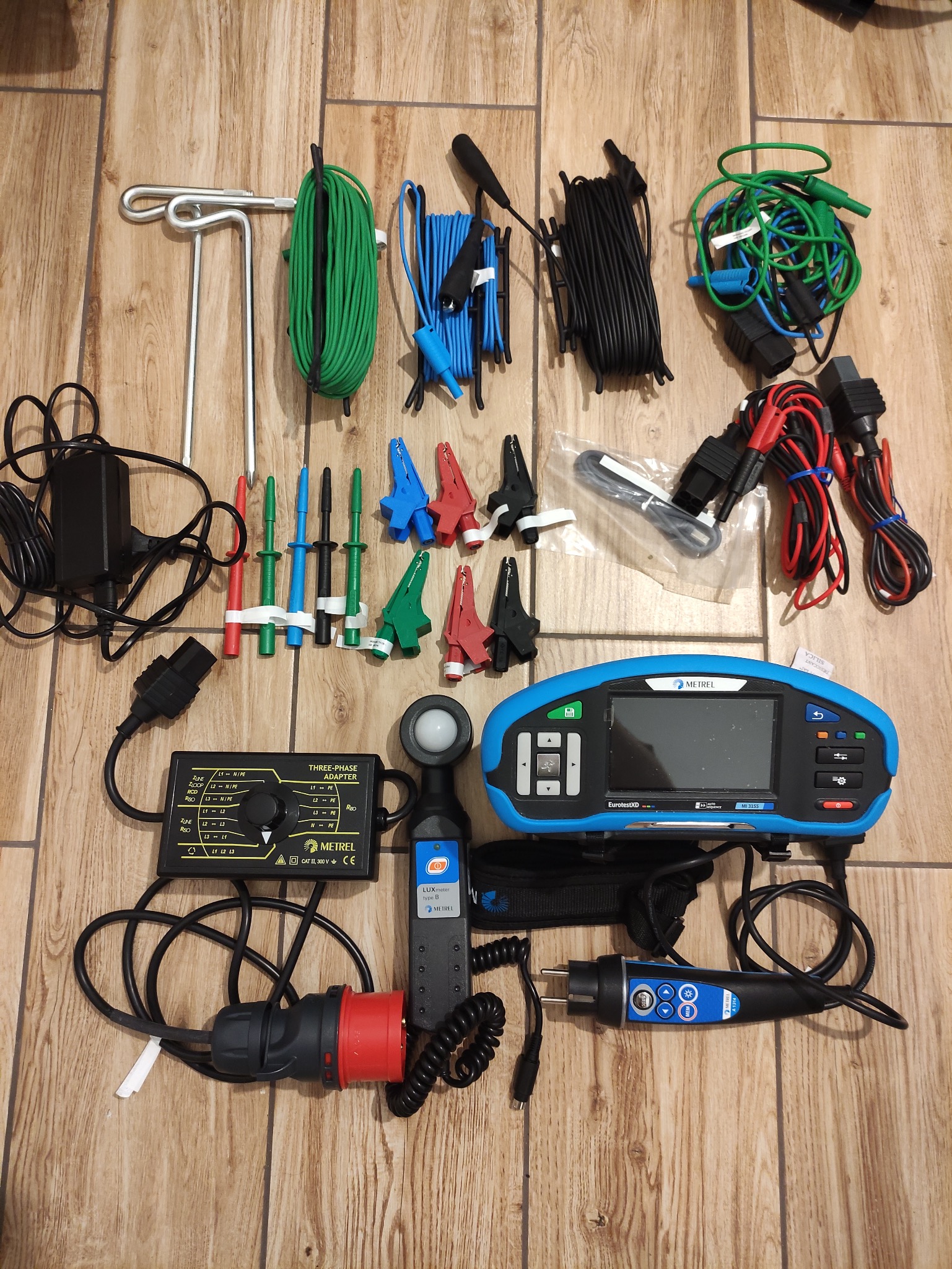 Zestaw narzędzi i akcesoriów elektrycznych Metrel na drewnianym podłożu. Widoczne mierniki, adaptery, przewody i sondy pomiarowe w różnych kolorach.