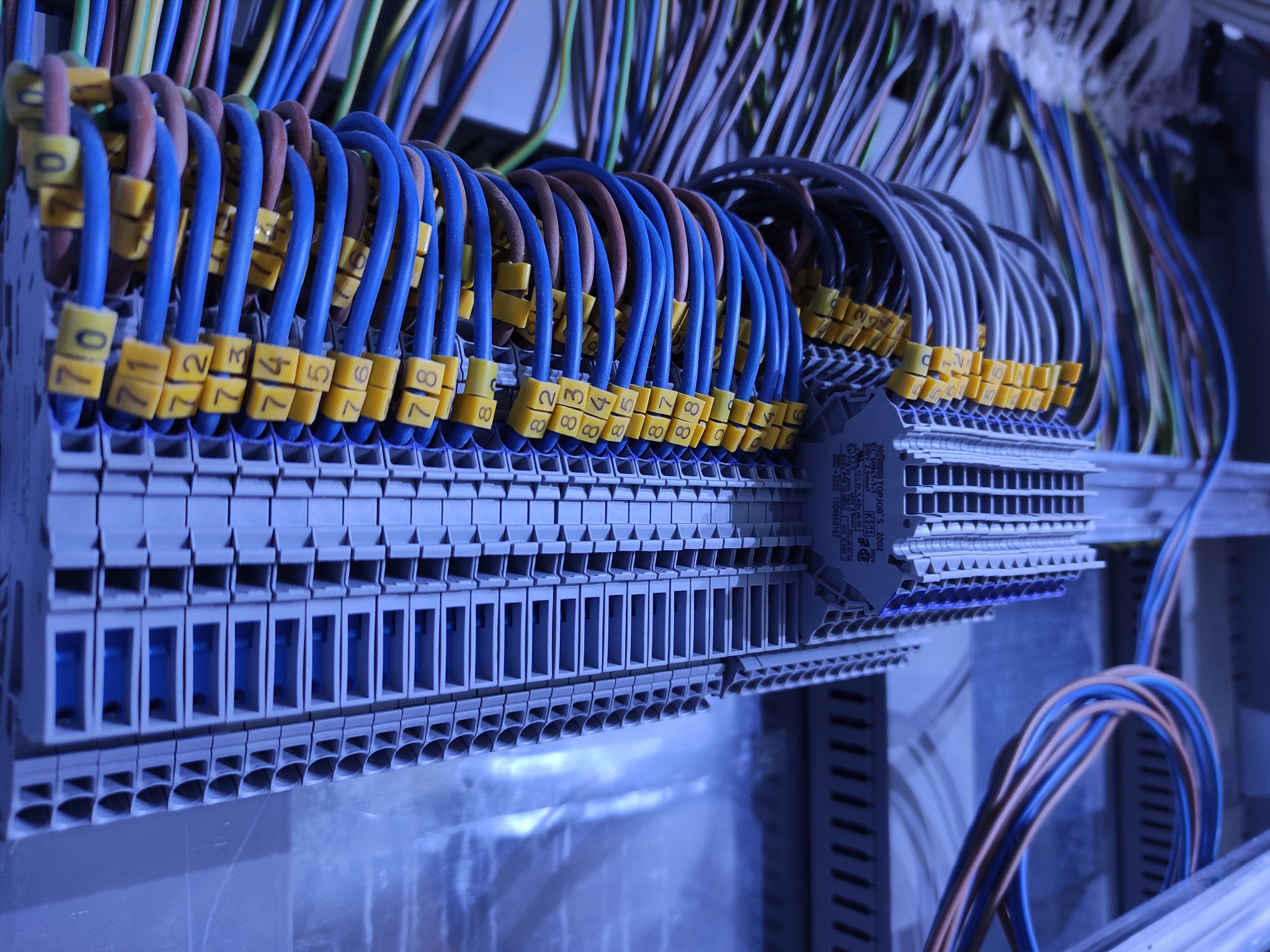 Szczegółowe ujęcie rozdzielnicy elektrycznej z podłączonymi, oznaczonymi przewodami w odcieniach niebieskiego, brązowego i zielonego, z żółtymi oznaczeniami numerów.