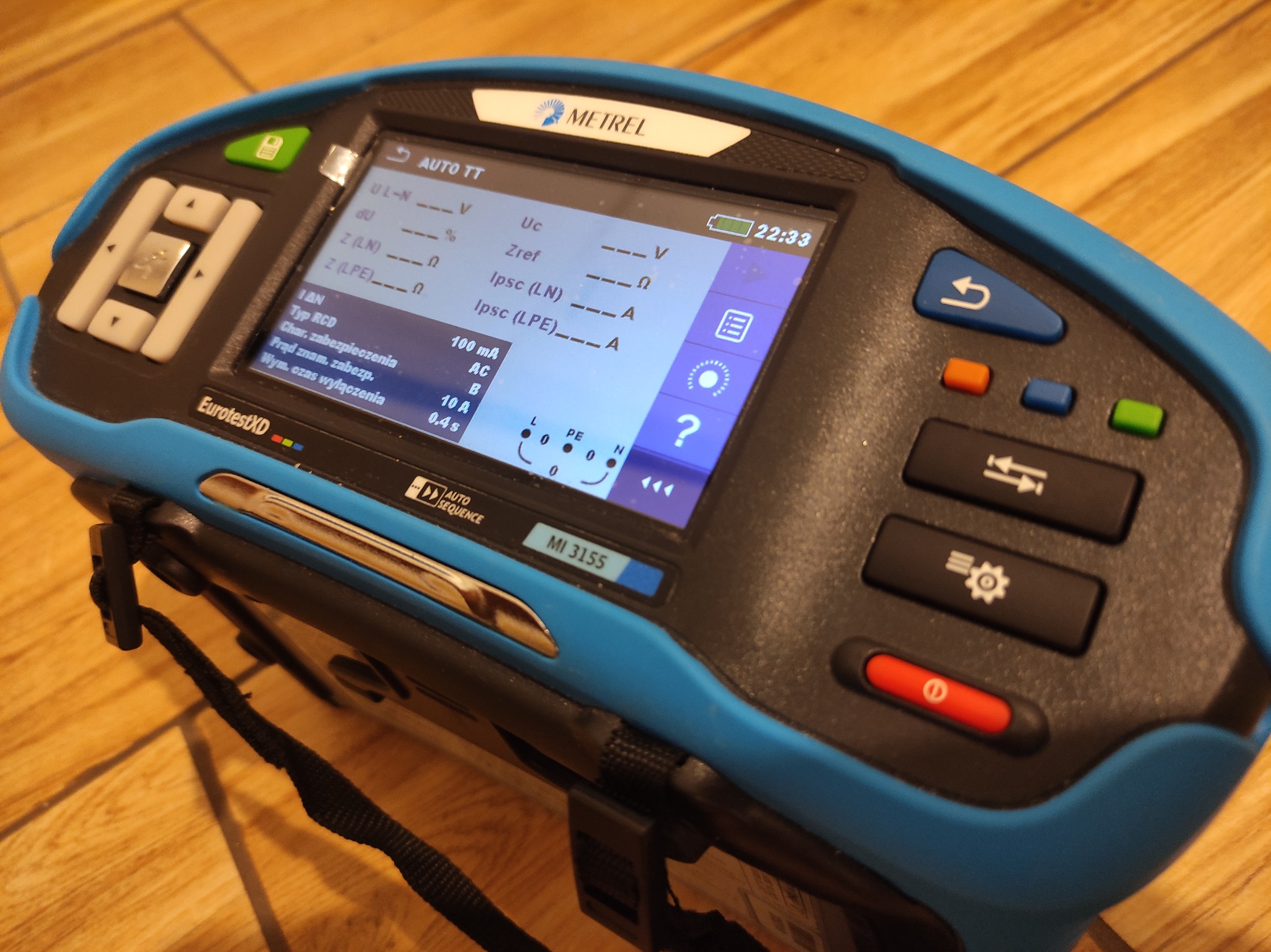 Miernik instalacji elektrycznych Metrel MI 3155 EurotestXD z wyświetlaczem parametrów, leżący na drewnianej powierzchni. Widoczne przyciski funkcyjne i pasek.