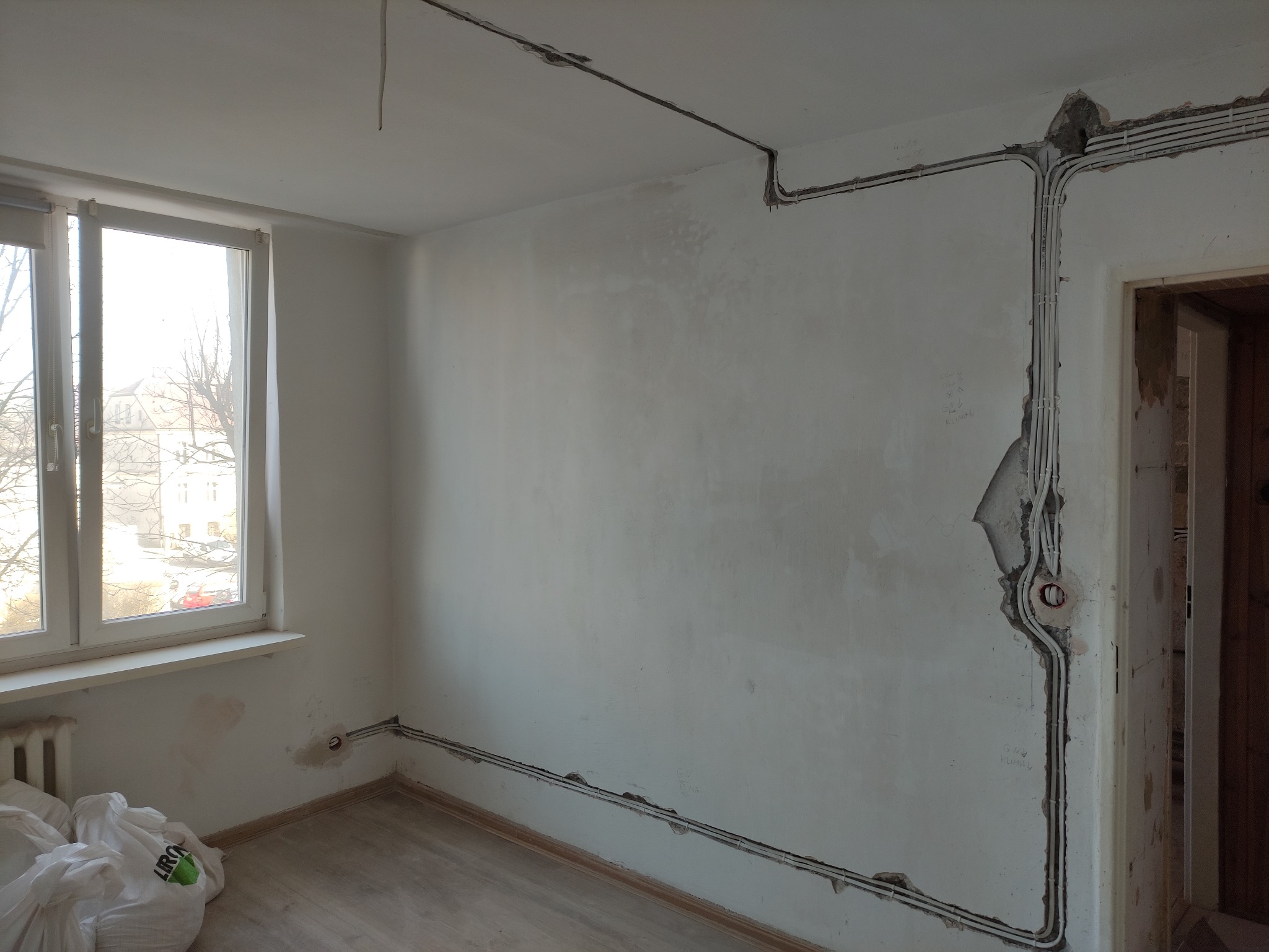 Białe kable elektryczne ułożone w bruzdach na ścianie w trakcie remontu pokoju. Widoczne puszki instalacyjne i fragmenty instalacji podtynkowej.