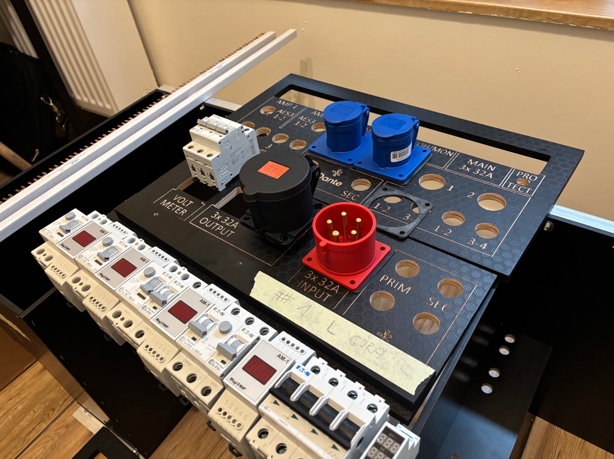 Rozdzielnica elektryczna z gniazdami 32A, bezpiecznikami EATON i miernikiem napięcia. Widoczne okablowanie i oznaczenia. Całość na czarnej płycie z otworami.
