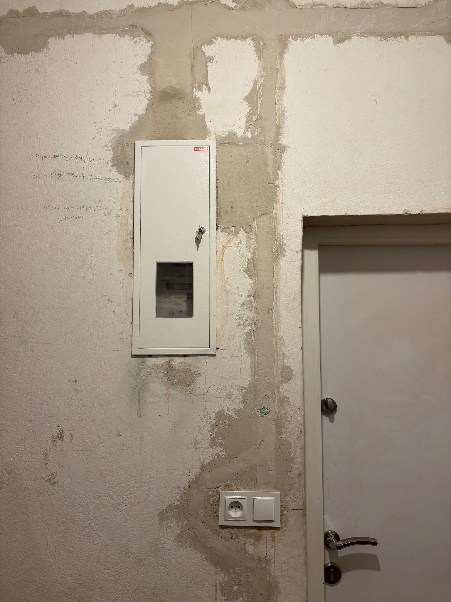 Biała skrzynka elektryczna z kluczykiem na surowej ścianie, obok gniazdko i drzwi. Widoczne ślady po remoncie i notatki na ścianie.