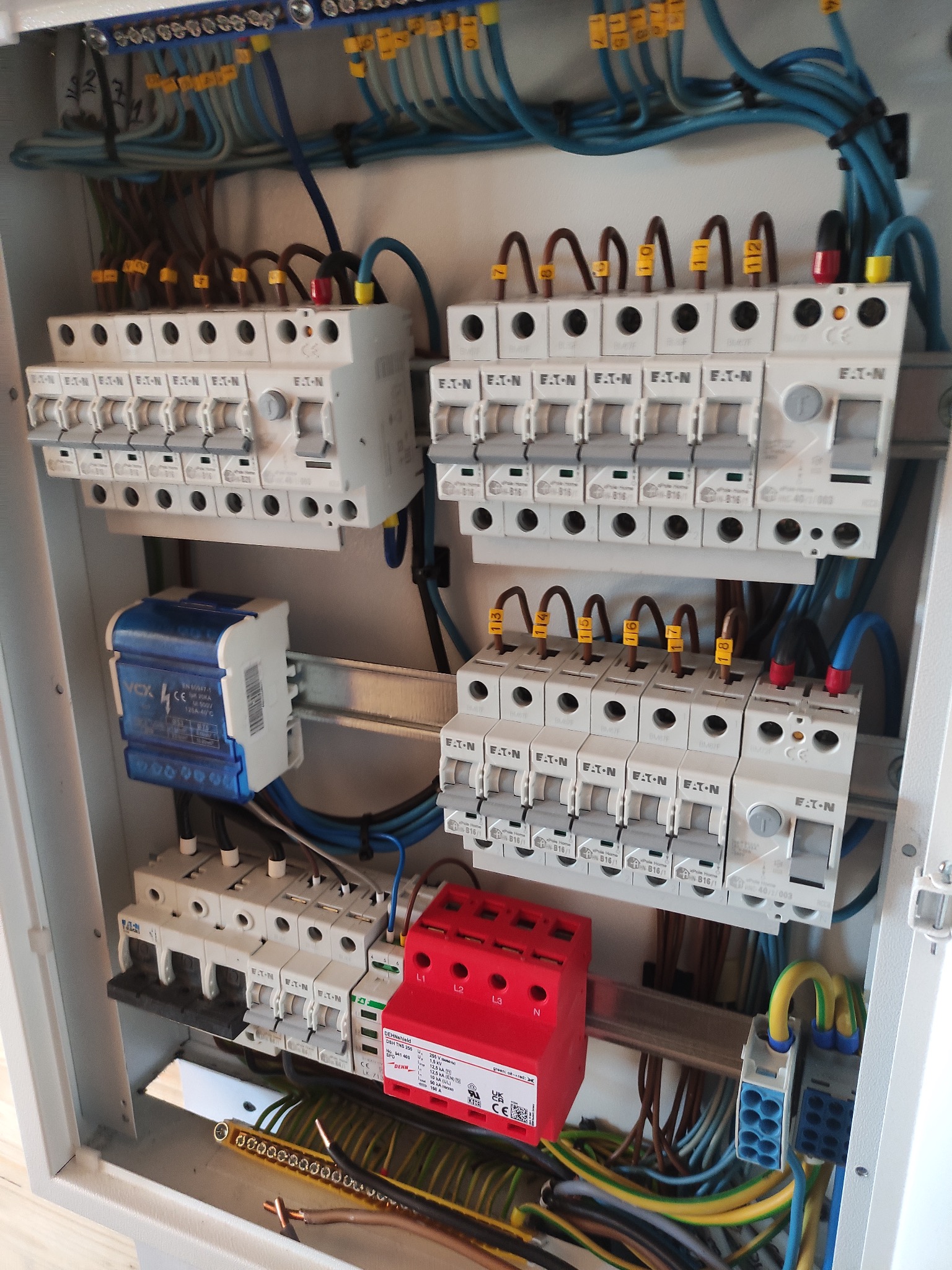 Rozdzielnica elektryczna z bezpiecznikami EATON, zabezpieczeniem przeciwprzepięciowym DEHN w kolorze czerwonym i niebieskim licznikiem VOX. Widoczne kable z oznaczeniami.