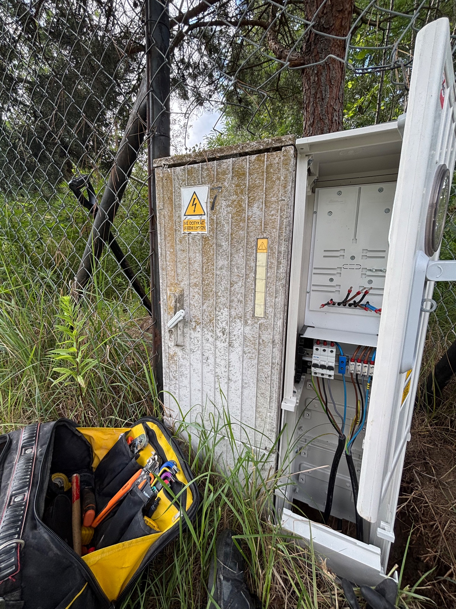 Otwarta skrzynka elektryczna z widocznymi przewodami i zabezpieczeniami, obok torba z narzędziami na tle trawy i ogrodzenia z siatki.